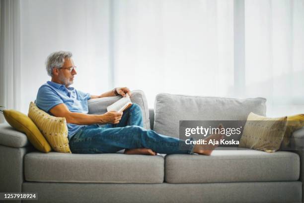 homme mûr lisant un livre sur le sofa. - comfortable photos et images de collection