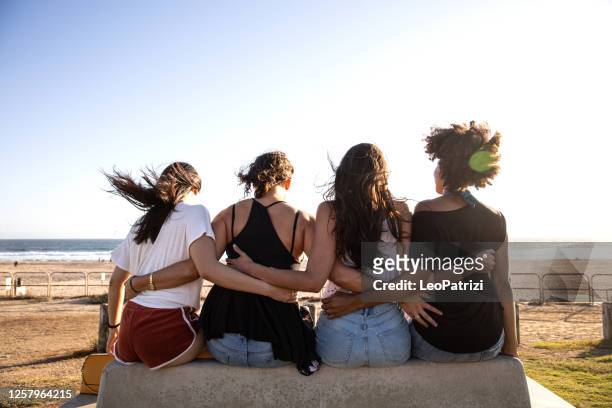 cuatro amigos abrazándose sentados en el banco frente a una playa - four people fotografías e imágenes de stock
