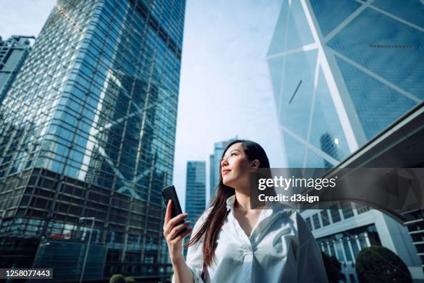 confidence and successful young asian businesswoman looking up to sky while using smartphone on the go in financial district, against corporate skyscrapers during the day in the city - china banking regulatory commission stockfoto's en -beelden