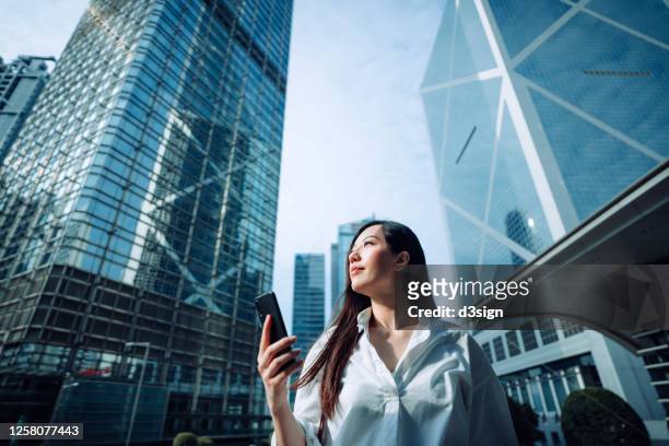 confidence and successful young asian businesswoman looking up to sky while using smartphone on the go in financial district, against corporate skyscrapers during the day in the city - hong kong advertising stock pictures, royalty-free photos & images