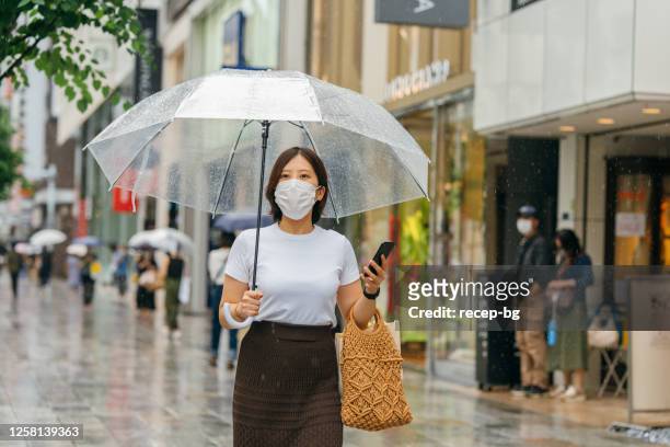 young woman holding umbrella and smart phone while walking in city during rain - rainy season stock pictures, royalty-free photos & images