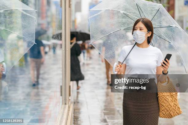 young woman holding umbrella and smart phone while walking in city during rain - rainy season stock pictures, royalty-free photos & images