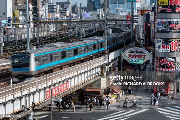 People wait to cross the street in front of the popular Ameyoko shopping district as a train passes by in the Ueno area of Tokyo on May 30, 2023.