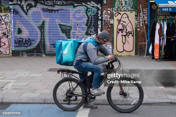 Deliveroo food delivery rider / cycle courier checks his smartphone on Whitechapel High Street on 25th May 2023 in London, United Kingdom. It acts as...