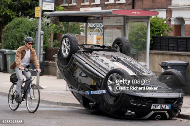 Cyclist passes the aftermath of a car crash in which an overturned Nissan hatchback remains at the side of the road between Brixton and Camberwell on...