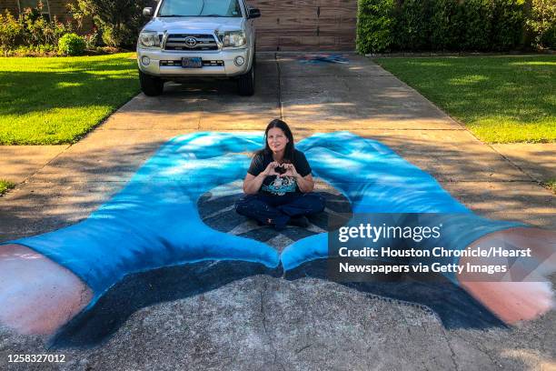 Artist Anat Ronen sits inside of a pair of hands wearing blue surgical gloves that she created as an anamorphic street painting with Tempera paint,...