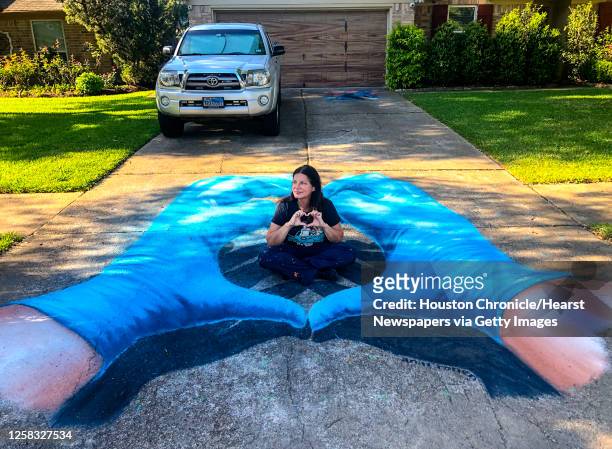 Artist Anat Ronen sits inside of a pair of hands wearing blue surgical gloves that she created as an anamorphic street painting with Tempera paint,...