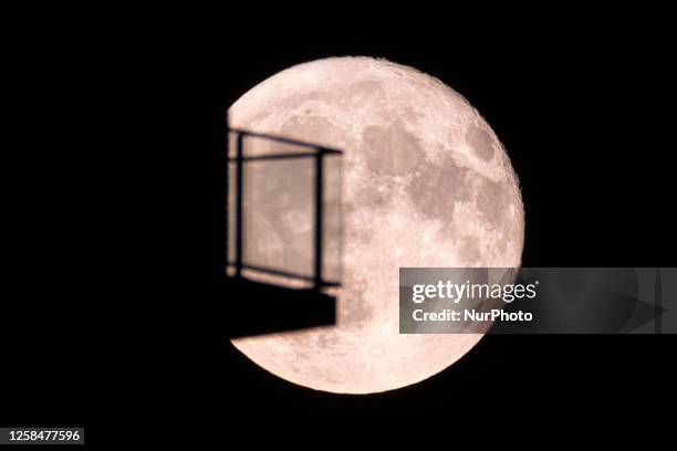Colorful pink Strawberry moon illuminates the night sky behind balconies from houses. Close-up of the June full moon, nicknamed as Strawberry moon as...