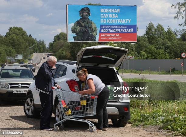 An elderly couple loads the trunk of their cars with purchases near a supermarket as a military propaganda billboard is seen in the background on...