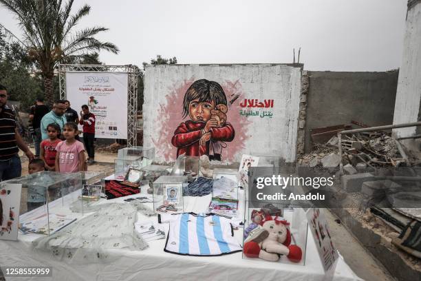 Palestinian kids stand near an exhibition of the clothes and toys of killed children and a 'Free Gaza' themed graffiti named 'Invasion Kills...