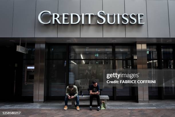 Two men sit under a sign of Credit Suisse displayed on a building in Lugano, on June 9, 2023. UBS is set to finalise the takeover of Credit Suisse on...