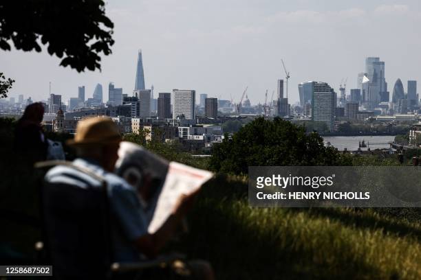 Man sits on a camping chair and reads the newspaper while enjoying the sun at Greenwich Park backdropped by the City business and financial district,...