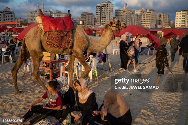 Youth leads a camel past beachgoers along a beach at sunset in Gaza City on June 14, 2023.