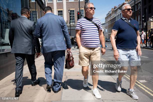 From the rear we see two men wearing impractical business suits walking past two others wearing cooler, more sensible shorts and casual wear for the...