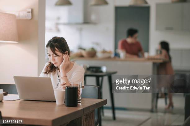 an asian chinese mid adult woman working in dining room typing using her laptop while her husband preparing food in the kitchen with their daughter - telecommuting couple stock pictures, royalty-free photos & images