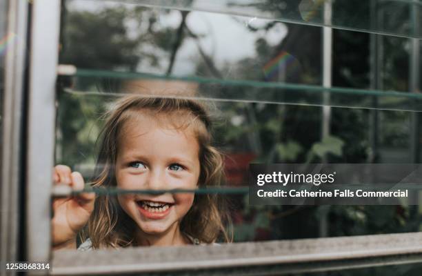 happy girl peeks through a slotted greenhouse window - girl looking through window stock pictures, royalty-free photos & images