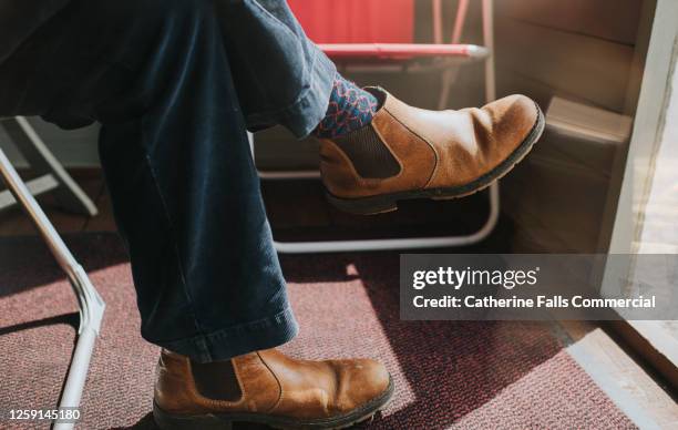 male wearing brown leather chelsea boots, legs crossed at the knee, sitting in a sun room. - brown shoe fotografías e imágenes de stock