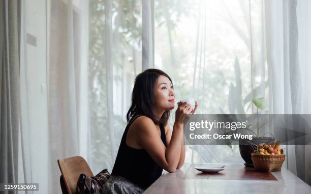 young woman drinking coffee near window at home - asian drink photos et images de collection