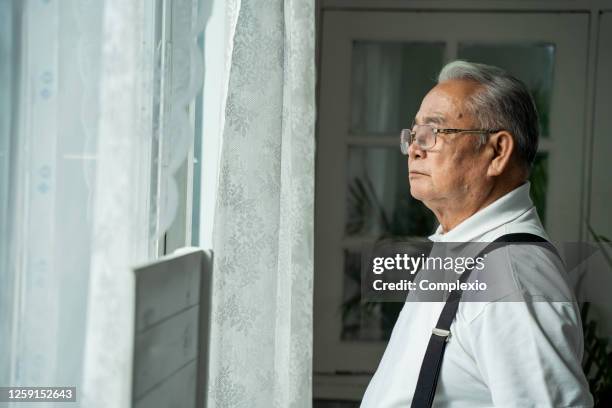 close up of pensive old man with glasses looking out of window feeling lonely. 70s asian pensioner missing children and wife. - widow pension stock pictures, royalty-free photos & images