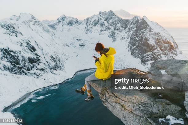 mujer sentada en el acantilado y usando el teléfono inteligente en la isla de lofoten en la nieve - winter landscape fotografías e imágenes de stock