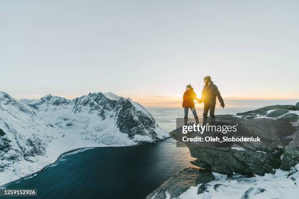 heterosexual couple standing on cliff and lookin at seaside of lofoten island in snow - winter norway stock pictures, royalty-free photos & images