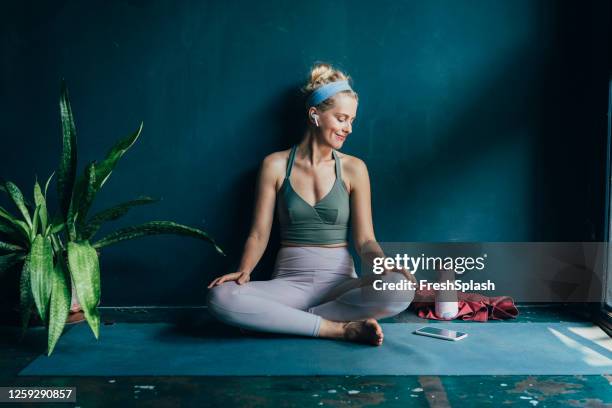 lächelnde blonde frau mit drahtlosen kopfhörern mit ihrem smartphone vor ihrem home workout - yoga stock-fotos und bilder