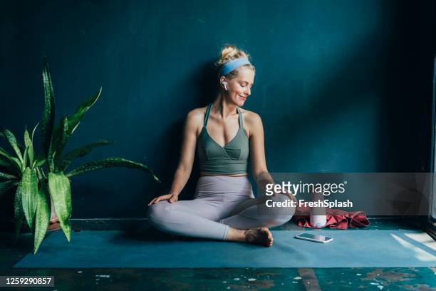 smiling blonde woman with wireless earphones using her smartphone before her home workout - yoga stock pictures, royalty-free photos & images