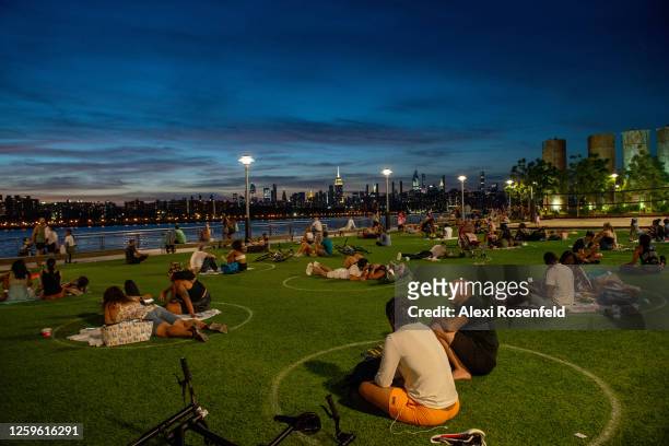 People with and without masks masks sit in circles to observe social distancing with the Manhattan skyline in the background in Domino Park as the...