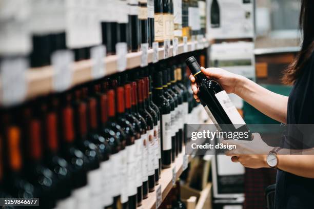 close up of young asian woman walking through supermarket aisle and choosing a bottle of red wine from the shelf in a supermarket - weinflaschen stock-fotos und bilder