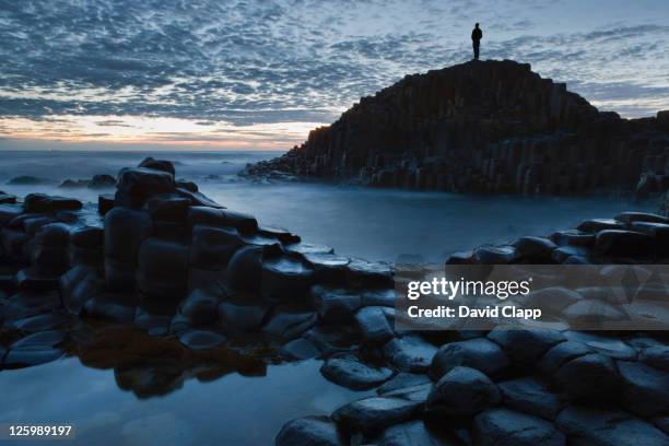 a lone figure stands on a basalt rock outcrop at the giants causeway at dusk, county antrim, northern ireland - rock formation stock pictures, royalty-free photos & images