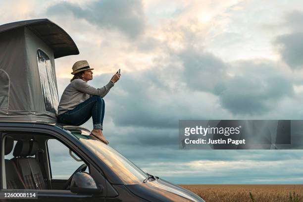 woman taking a photo whilst sitting on roof of camper at sunset - reise stock-fotos und bilder