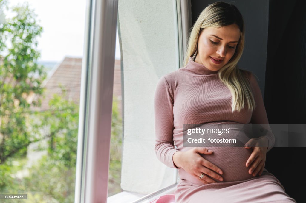 Woman feeling her baby moving in her belly while relaxing on the window sill