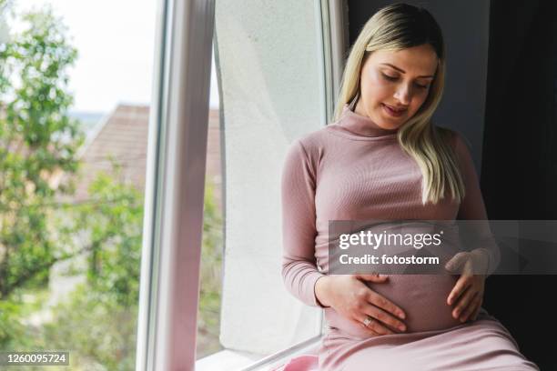 woman feeling her baby moving in her belly while relaxing on the window sill - pink dress stock pictures, royalty-free photos & images