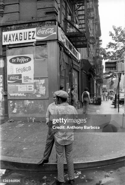 View of two men standing on an unidentified Harlem street corner, New York, June 1975. Other figures are visible standing outside a storefront,...