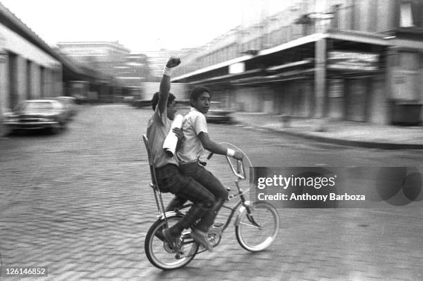 View of two boys riding a bicycle along an unidentified Harlem street, with the passenger raising his fist in salute, New York, June 1972.