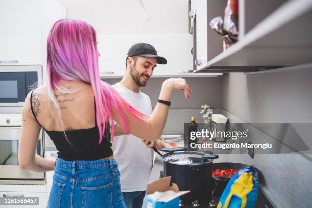 young woman with long pink hair and bearded man wearing baseball cap standing in a kitchen, cooking. - baseball tattoo photos et images de collection