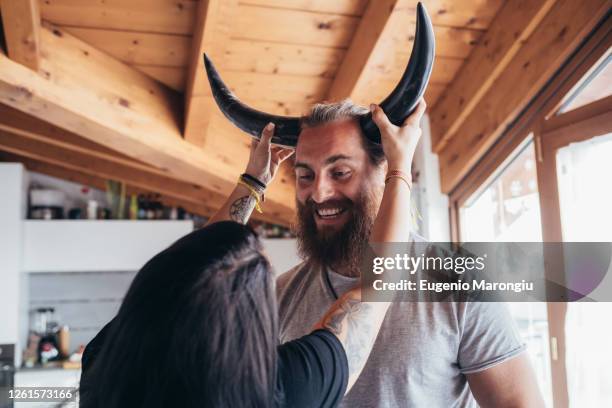 woman holding cow horns onto head of smiling bearded man with brunette hair. - horny couple stock pictures, royalty-free photos & images