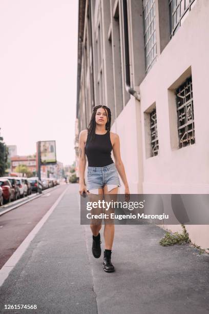 portrait of young woman with dark brown hair, wearing black vest and denim shorts, walking down street. - shorts down stock pictures, royalty-free photos & images