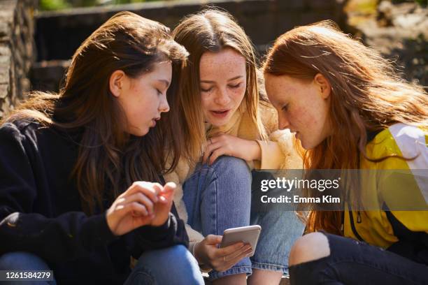 three teenage girls sitting outdoors, checking their mobile phones. - 3 teenagers mobile outdoors stockfoto's en -beelden