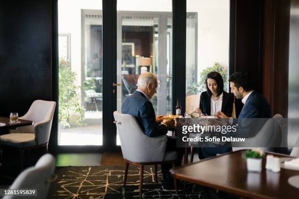 two businessmen and woman having working lunch in hotel restaurant - business lunch stock-fotos und bilder