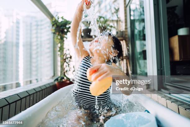 joyful little asian girl playing with rubber duck and toy watering can having fun playing in wading pool in the balcony at home, splashing water all over her face - asian water splash stock pictures, royalty-free photos & images