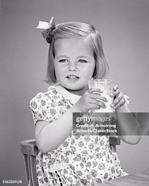 1940s Little Blonde Girl Bow In Her Hair Print Dress Drinking A Glass Of Milk Sitting At Child'S Table And Chair