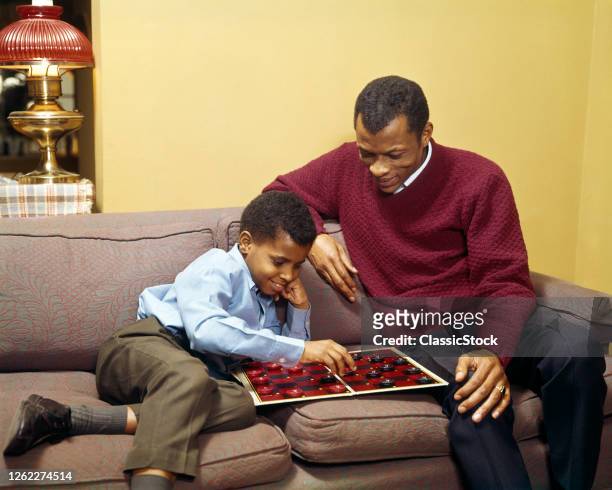 1960s 1970s African American Father And Son Sitting On Couch Together In Living Room At Home Playing A Game Of Checkers