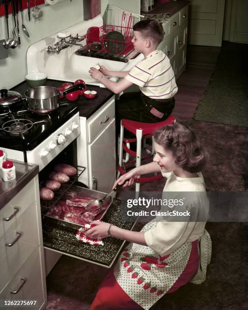 1950s Kitchen Smiling Teenage Girl Sister Kneeling by Oven Roasting Chops Baking Potatoes Boy Brother Washing Dishes At Sink