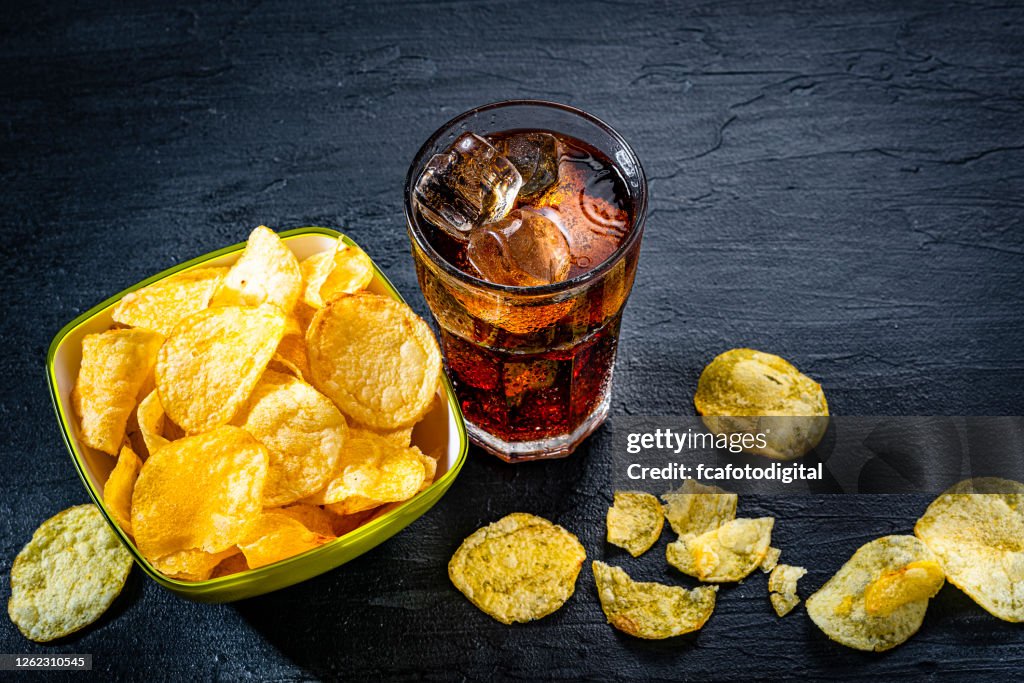 Cola soda glass and potato chips in a bowl shot on black slate table
