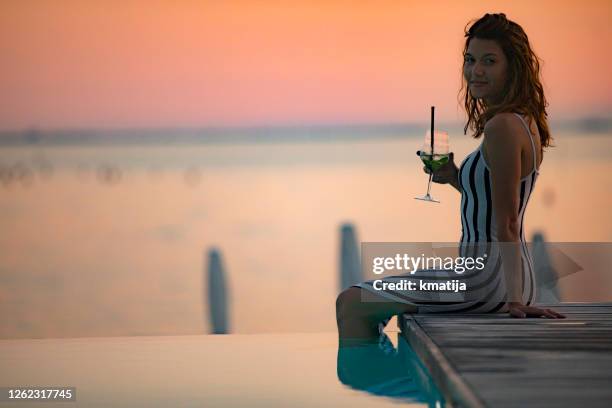 junge frau trinken cocktail sitzen deiner pool in der dämmerung - cocktail sonnenuntergang stock-fotos und bilder