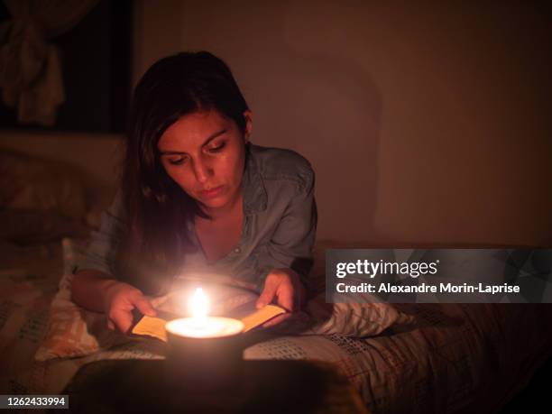 young hispanic woman reads a book lying in a bed on a leaf pattern pillow at night with a candle - kerzenschein stock-fotos und bilder