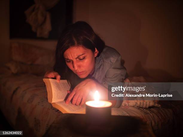 young hispanic woman reads a book lying in a bed on a leaf pattern pillow at night with a candle - hot latin nights stock pictures, royalty-free photos & images