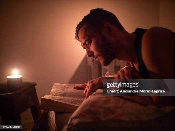 young caucasian man reads a book in bed by candlelight - candlelight stock pictures, royalty-free photos & images