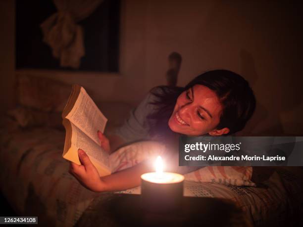 young hispanic woman reads a book lying in a bed on a leaf pattern pillow at night with a candle - hot latin nights stock pictures, royalty-free photos & images